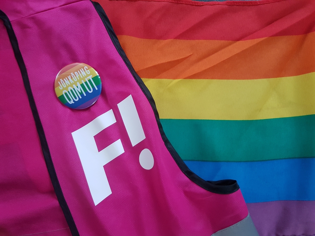 F!-logga på rosa väst i bakgrunden en regnbågsflagga